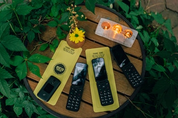 Cuatro teléfonos móviles antiguos sobre dos paneles amarillos 'Phone Zone' en una mesa de madera con hojas, una flor amarilla y tres velas encendidas
