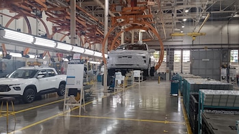Vista interior de una planta automotriz mostrando un vehículo camuflado suspendido en un transportador y una camioneta blanca en el suelo