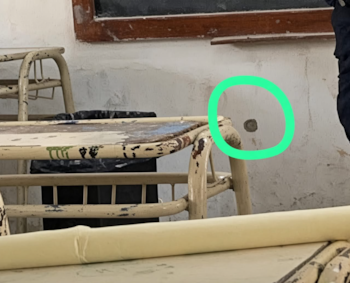 Un orificio en la pared marcado con un círculo verde dentro de un aula con bancos de madera y pizarrón