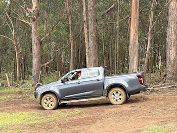 Vista lateral de un camión Isuzu D-Max 4x4 gris oscuro conduciendo en un sendero de tierra embarrado, con un conductor visible y un bosque de árboles altos al fondo