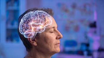 Perfil de una persona con los ojos cerrados y una proyección digital traslúcida de un cerebro humano iluminado en su interior.