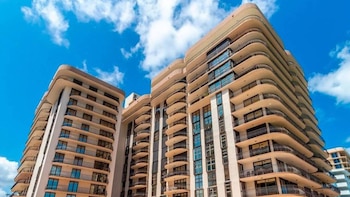 Edificio de gran altura color claro con balcones semicirculares y ventanas de vidrio oscuro bajo un cielo azul con nubes blancas. Vista desde abajo