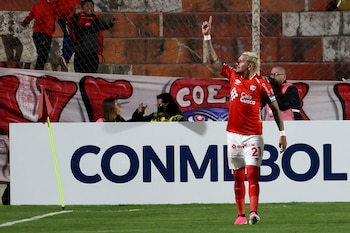 Jugador de fútbol con camiseta roja y pantalón blanco en un campo verde, levanta el brazo derecho. Un cartel de CONMEBOL y un banderín de córner