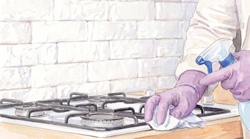 Una mano con guante morado sostiene un paño blanco sobre una estufa de gas de cuatro quemadores con rejillas negras. Hay un rociador azul y una pared de ladrillos blancos.