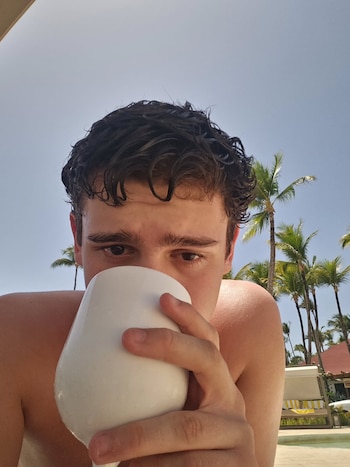 Primer plano de un joven bebiendo de una taza blanca. Tiene cabello oscuro mojado. Al fondo se ven palmeras y un cielo azul