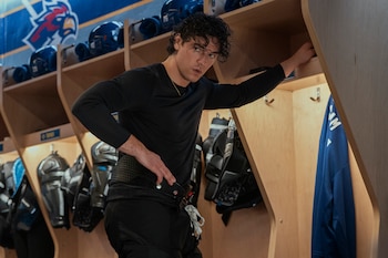 Hombre joven de cabello rizado con camiseta y pantalones negros posa en un vestuario. Se ven casilleros de madera, cascos y equipo de hockey, y un logo de ave azul al fondo