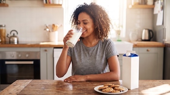 Una mujer de cabello rizado bebe leche de un vaso transparente junto a un cartón de leche y un plato de galletas sobre una mesa.