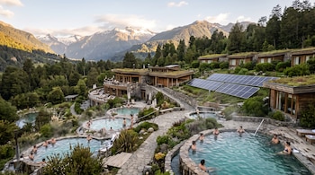 Piscinas de agua termal con personas, edificaciones de madera con paneles solares y vegetación frente a montañas nevadas.