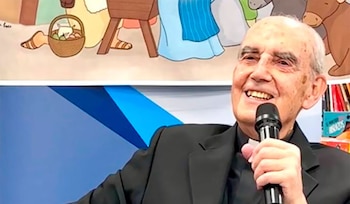 Un hombre con traje negro y alzacuello sostiene un micrófono frente a una lámina con personajes de una ilustración infantil