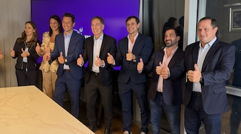 Dos hombres sonrientes, Diego Santilli y Adrián Núñez, con trajes, posan con el pulgar en alto frente a una pantalla morada con el logo de La Libertad Avanza