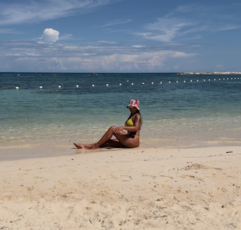 Una mujer con traje de baño amarillo, sombrero colorido y gafas de sol está sentada en la orilla de una playa frente al mar
