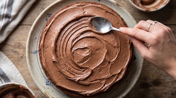 Plano cenital de una mano que aplica crema de chocolate con una cuchara sobre una torta redonda.
