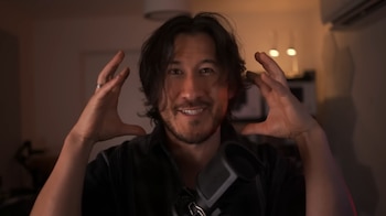 Un hombre de cabello castaño oscuro con barba y camisa oscura gesticula con ambas manos frente a un micrófono; se ven dos lámparas y un aire acondicionado detrás