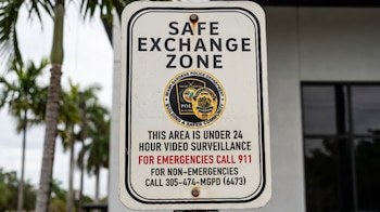 Un cartel blanco y negro con la leyenda 'SAFE EXCHANGE ZONE' del Departamento de Policía de Miami Gardens, con palmeras en el fondo borroso