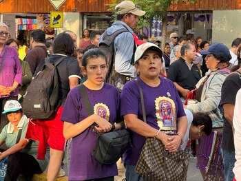 Dos mujeres con camisetas moradas y estampados religiosos permanecen de pie en medio de una multitud en una calle urbana