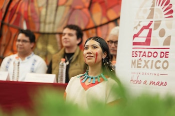 Una mujer con pintura facial, collar tradicional y traje claro permanece ante un panel publicitario del Estado de México