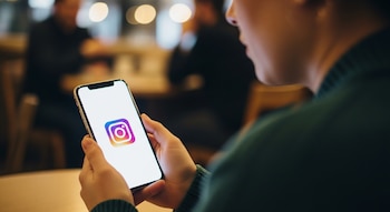 Plano medio de una persona de perfil, sosteniendo un smartphone con ambas manos, cuya pantalla iluminada muestra prominentemente el logo colorido de Instagram.