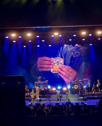 Banda tocando en un escenario con luces azules. Detrás, una pantalla gigante muestra manos sosteniendo cartas Pokémon junto a un micrófono