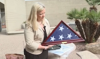 Una mujer rubia con chaqueta clara sostiene una vitrina triangular de madera que contiene una bandera de Estados Unidos plegada