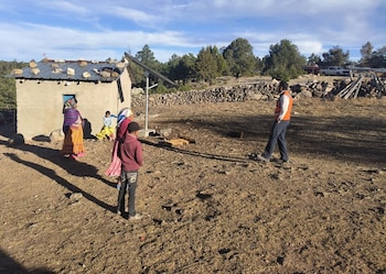 Un panel solar junto a una casa de color claro en un terreno árido. Varias personas, incluyendo niños, están paradas frente a la casa. Al fondo hay árboles y una pared de piedra