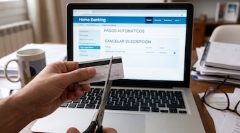 Primer plano de una mano sosteniendo una tarjeta de crédito, con tijeras a punto de cortarla. Al fondo, un portátil muestra la pantalla de Home Banking con la opción de cancelar suscripciones.