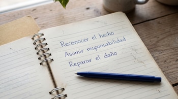 Libreta de espiral abierta con las frases Reconocer el hecho, Asumir responsabilidad y Reparar el daño escritas en azul junto a un bolígrafo.