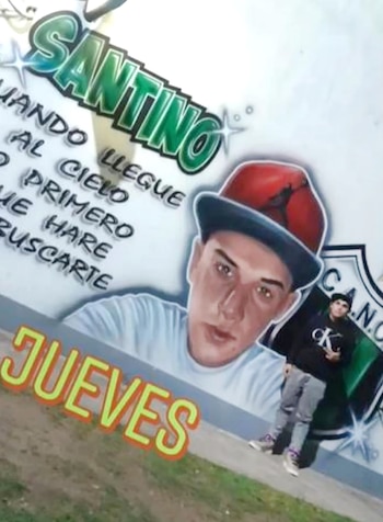 Mural en pared blanca con retrato aerografiado de joven con gorra, texto "SANTINO", y un joven de pie en ropa oscura