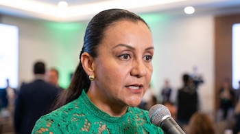 Primer plano de la diputada Gabriela Benavides Cobos del Partido Verde hablando a un micrófono. Viste una blusa verde de encaje y aretes dorados. El fondo está borroso con personas y luces.