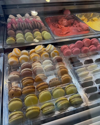 Vitrina con bandejas de macarons rosados, verdes, amarillos y marrones. Dos recipientes con helado rojo y amarillo. Bandejas de bombones de chocolate claro y oscuro