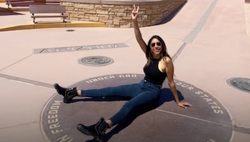 Mujer sentada sobre una placa circular grabada que dice "FOUR STATES" y "ARIZONA", con jeans, top negro y botas, sonriendo y haciendo el signo de la paz