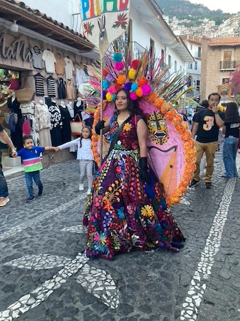 Mujer con vestido bordado, tocado de pompones y estandarte rosa con cara de jaguar desfila por calle empedrada. Se ven personas observando