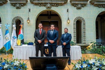 Tres hombres con traje formal posan de pie junto a banderas de Guatemala en un patio con arcos de piedra y arreglos florales