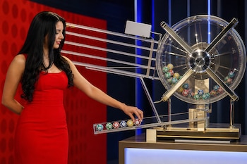 Una mujer con vestido rojo junto a un bombo de lotería transparente que contiene bolas de colores numeradas sobre una bandeja.