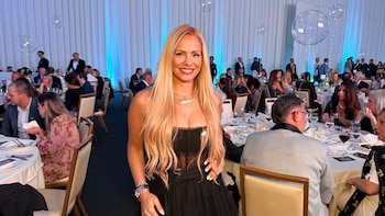 Antonella Fredianelli con cabello rubio y vestido negro sonríe frente a mesas de gala con invitados y globos claros en una sala de eventos