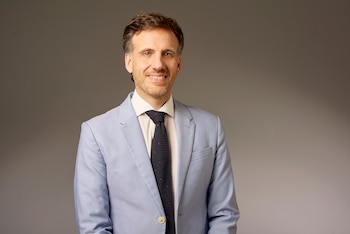 Jorge Dotto sonríe vestiendo un saco azul claro, camisa blanca y corbata oscura sobre un fondo gris de estudio