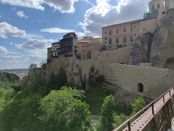 Edificios históricos sobre un acantilado de piedra, una barandilla metálica en primer plano, árboles en la base y un cielo con nubes