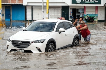 Tres hombres empujan un automóvil blanco varado en una calle urbana completamente cubierta por agua turbia frente a locales comerciales cerrados