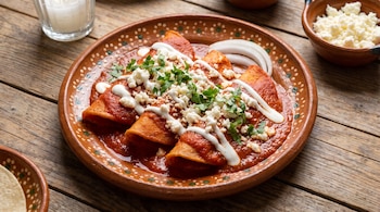 Tres entomatadas en salsa roja con crema, queso desmoronado, cilantro y cebolla sobre un plato de barro en una mesa de madera.