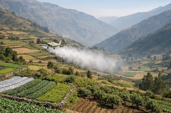 Un avión amarillo y azul dispersa una nube de humo blanco sobre campos de cultivo en terrazas, con montañas y un pequeño pueblo al fondo.
