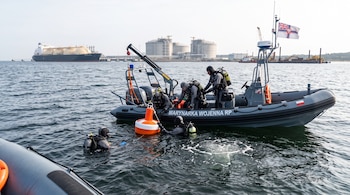 Buzos con trajes de neopreno en una lancha neumática y en el agua junto a una boya, con un buque y tanques de gas en el horizonte.