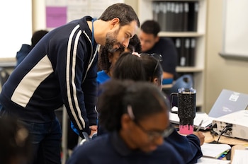 Un profesor barbudo se inclina sobre el escritorio de un estudiante en un aula, observando su trabajo. Otros estudiantes y un proyector son visibles