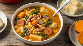 Primer plano de un tazón de cachupa con maíz, frijoles, carne, chorizo, yuca y camote, acompañado de yuca cocida y camote en rodajas.
