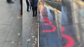 Panel metálico con nombres grabados y marcas de pintura roja en aerosol, junto a varias personas de pie sobre una acera de losas de piedra