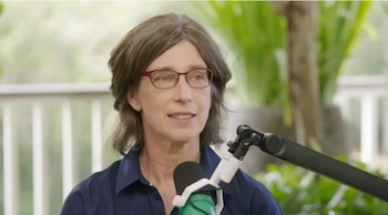 Mujer de mediana edad con cabello castaño, gafas rojas y camisa azul oscuro hablando en un micrófono. Fondo verde borroso con barandilla