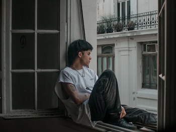 Joven sentado en un alféizar de ventana. Lleva camiseta blanca, pantalón oscuro, collar y arete. Edificio con balcón al fondo