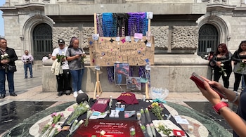 Personas de pie junto a un altar en la vía pública con carteles, fotografías, flores blancas y velas.