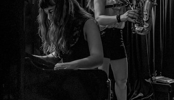 Imagen en blanco y negro de dos mujeres en un escenario; una está frente a una consola y la otra sostiene un saxofón, con un mezclador visible