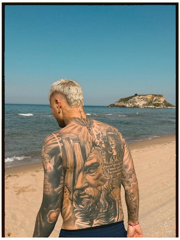 Hombre de espaldas con tatuajes grandes y elaborados en toda la espalda y brazos, de pie en una playa. El mar y una pequeña isla rocosa están al fondo bajo cielo azul
