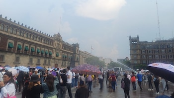 Vista de una multitud de personas con paraguas en una plaza mojada frente al Palacio Nacional. Otros edificios históricos y un chorro de agua son visibles bajo cielo nublado