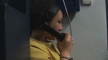 Una mujer con uniforme amarillo y esposas habla por un teléfono negro en una cabina oscura frente a una pared clara